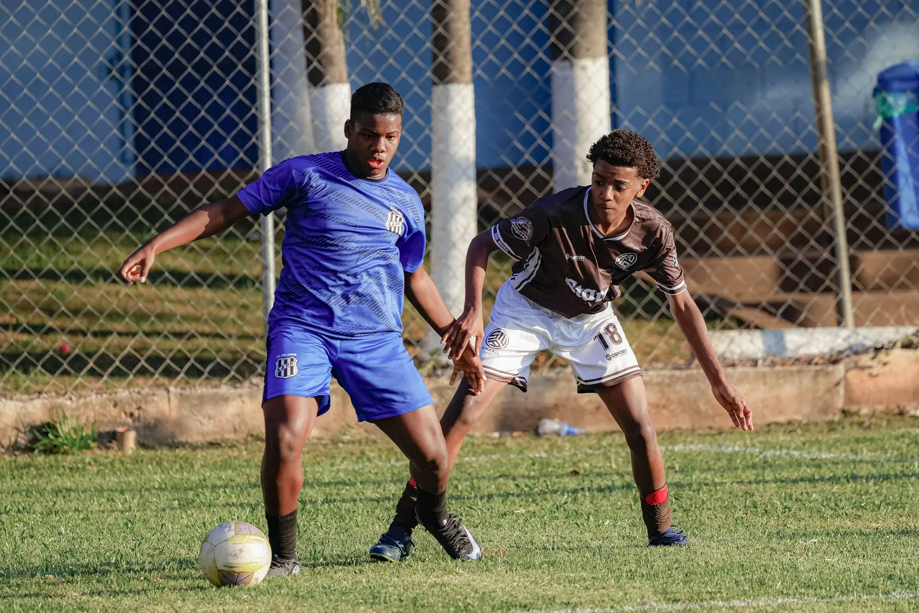Disputa de bola entre um atleta da SNA Brasil, de uniforme marrom, e um adversário de azul