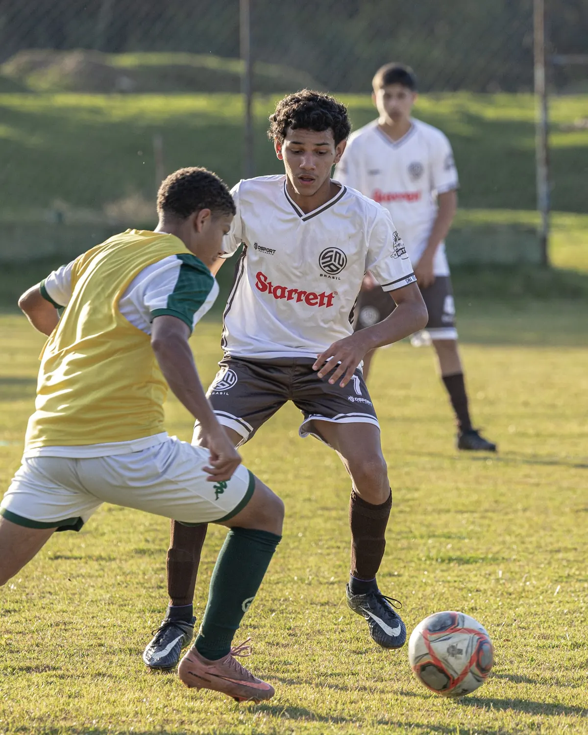 Atleta de uniforme branco protege a bola de um adversário de colete amarelo